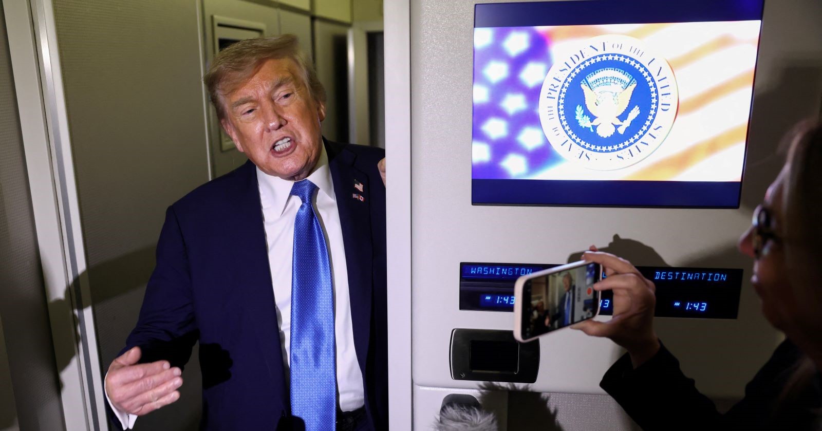 Donald Trump talks to media representatives on a plane, next to him a screen with the presidential seal of the USA. A person films him with his smartphone.